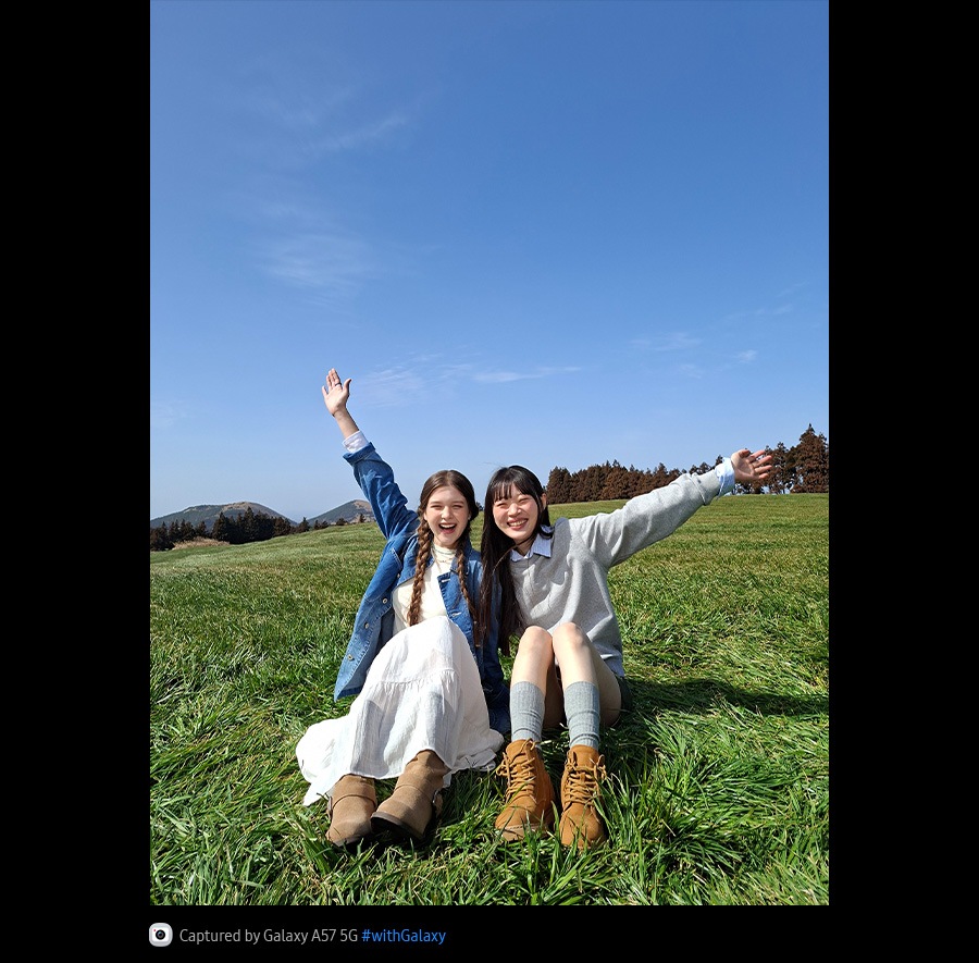 Fotografie cu două tinere așezate în mijlocul unui câmp deschis, cu iarbă pe fundalul cerului albastru, realizată cu camera foto cu unghi larg a modelului Galaxy A57 5G. Detaliile fotografiei sunt luminoase și intense, cu o calitate îmbunătățită. În partea de jos se află pictograma camerei foto, cu textul: „Capturat cu Galaxy A57 5G, hashtag cu Galaxy”.