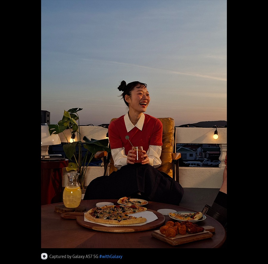 Fotografie cu o tanara zambitoare, pe o terasa in aer liber, realizata intr-un mediu slab iluminat, cu Galaxy A57 5G. Detaliile fotografiei sunt luminoase si clare. In partea de jos se afla pictograma camerei foto, cu textul: „Capturat cu Galaxy A57 5G, hashtag cu Galaxy”.