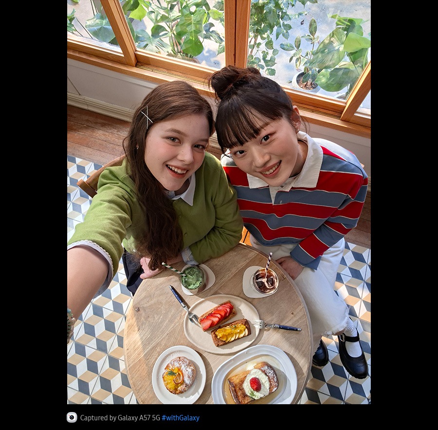 Fotografie selfie cu două tinere luând prânzul într-o cafenea luminată puternic, realizată de sus, cu camera foto frontală a modelului Galaxy A57 5G. Detaliile fotografiei sunt vibrante și colorate datorită Super HDR. În partea de jos se află pictograma camerei foto, cu textul: „Capturat cu Galaxy A57 5G, hashtag cu Galaxy”.