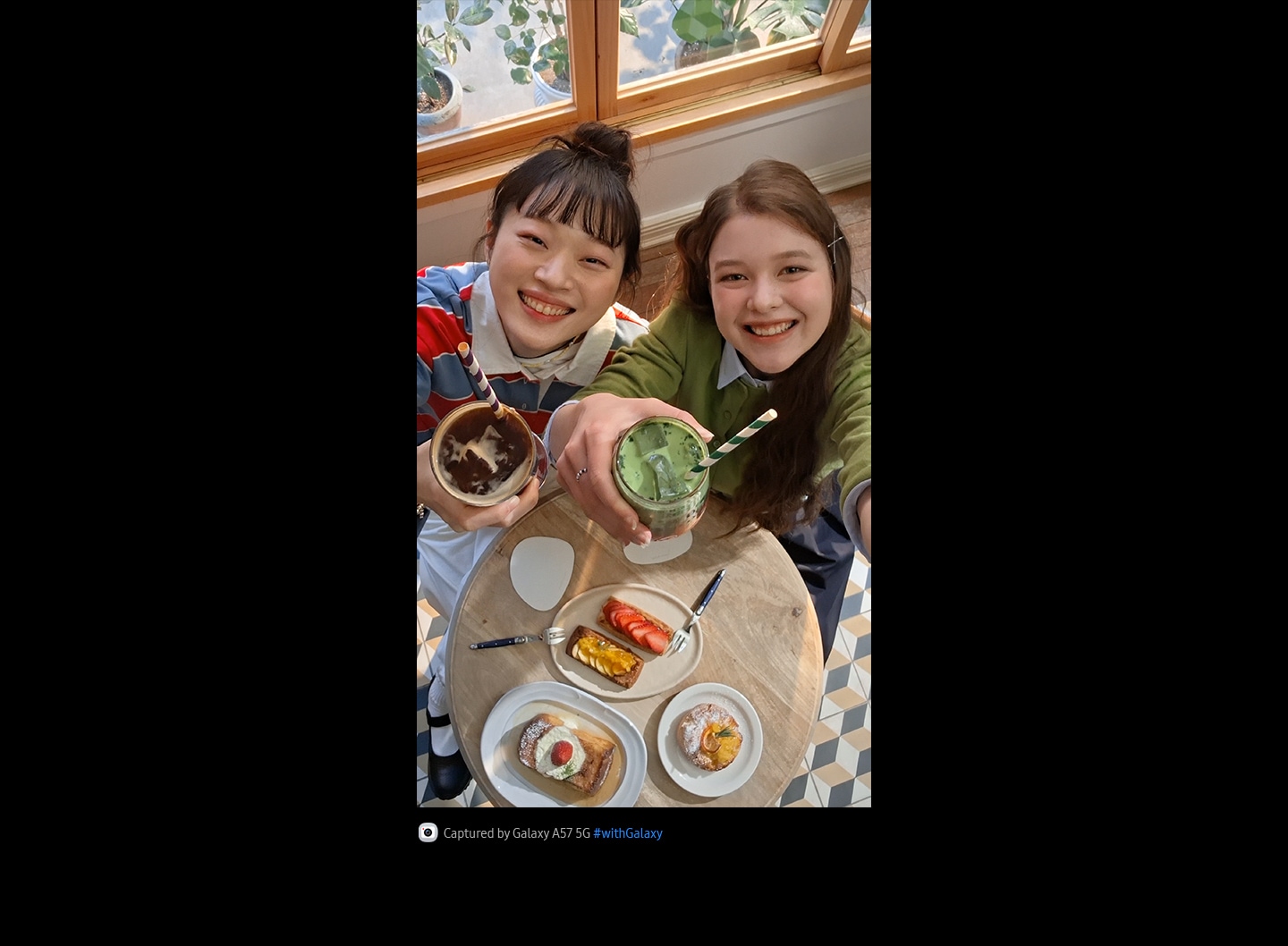 Videoclip selfie cu două tinere luând prânzul într-o cafenea luminată puternic, realizat de sus, cu camera foto frontală a modelului Galaxy A57 5G. Detaliile videoclipului sunt vibrante și colorate datorită Super HDR. În partea de jos se află pictograma camerei foto, cu textul: „Capturat cu Galaxy A57 5G, hashtag cu Galaxy”.
