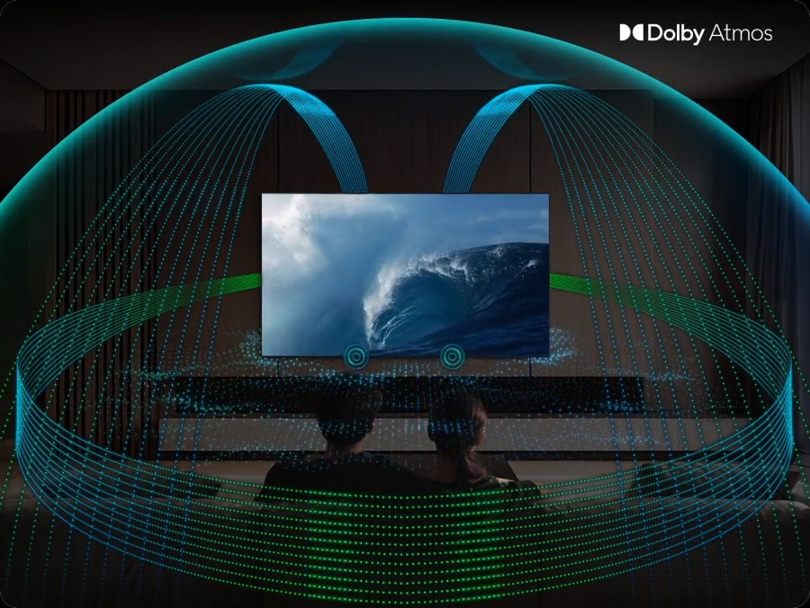 In a living room, a couple sits on a sofa and watches TV. The TV’s front speakers are lit up and soundwaves emanate from a TV’s side and up-firing speakers, travelling in multiple directions to surround the couple in a dome of sound and fill the room. Dolby Atmos logo.