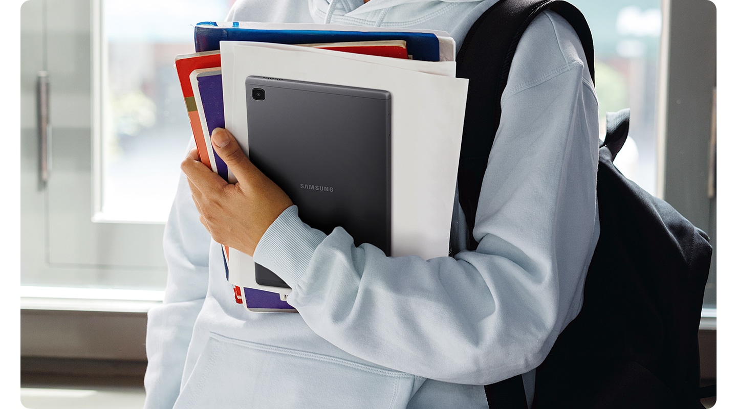 A person wearing a backpack and holding a pile of papers and textbooks. At the front of the pile is Galaxy Tab A7 Lite.