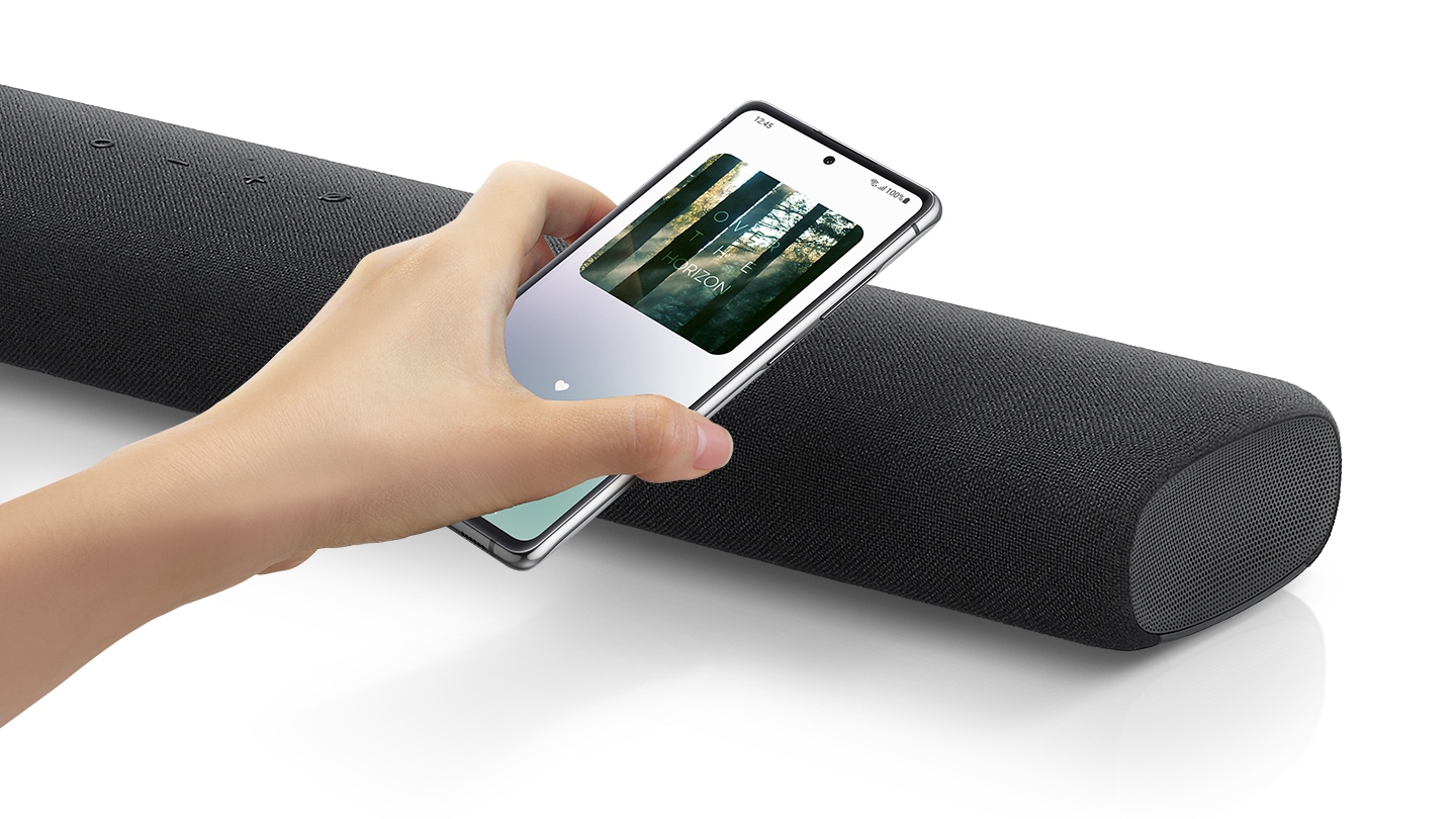 A hand taps a smartphone with the Samsung music app on-screen on the soundbar and the soundbar instantly plays music, showing how easy it is to switch from smartphone to soundbar.