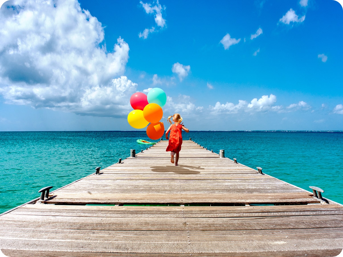 A girl is running on a dock holding a bundle of balloons. With Ultra Wide Camera, you can see more of the dock and the ocean.