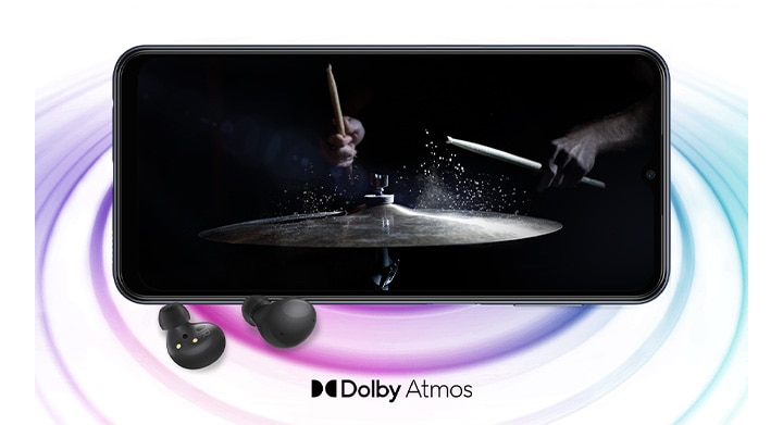 Galaxy M33 5G в ландшафтном режиме и изображение с человеком, играющим на барабанах на черном фоне на экране. Пара черных Galaxy Buds2 расположена перед устройством. Справа внизу находится логотип Dolby Atmos.