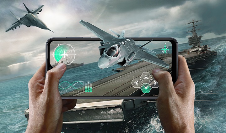 Две руки держат устройство Galaxy M33 5G горизонтально, играя в игру fps на истребителе. На заднем плане изображена внутриигровая атмосфера морского боя с большим авианосцем, вытянутым за пределы экрана. Изнутри экрана самолет-истребитель слегка выступает за пределы экрана. Вверху в тексте указаны температура, объем памяти и время автономной работы.