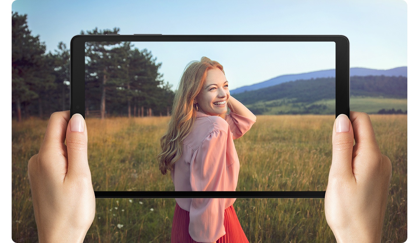 Руки держат Galaxy A7 Lite. На экране - женщина, стоящая в поле. Фотография выходит за границы экрана, чтобы представить широкий дисплей.