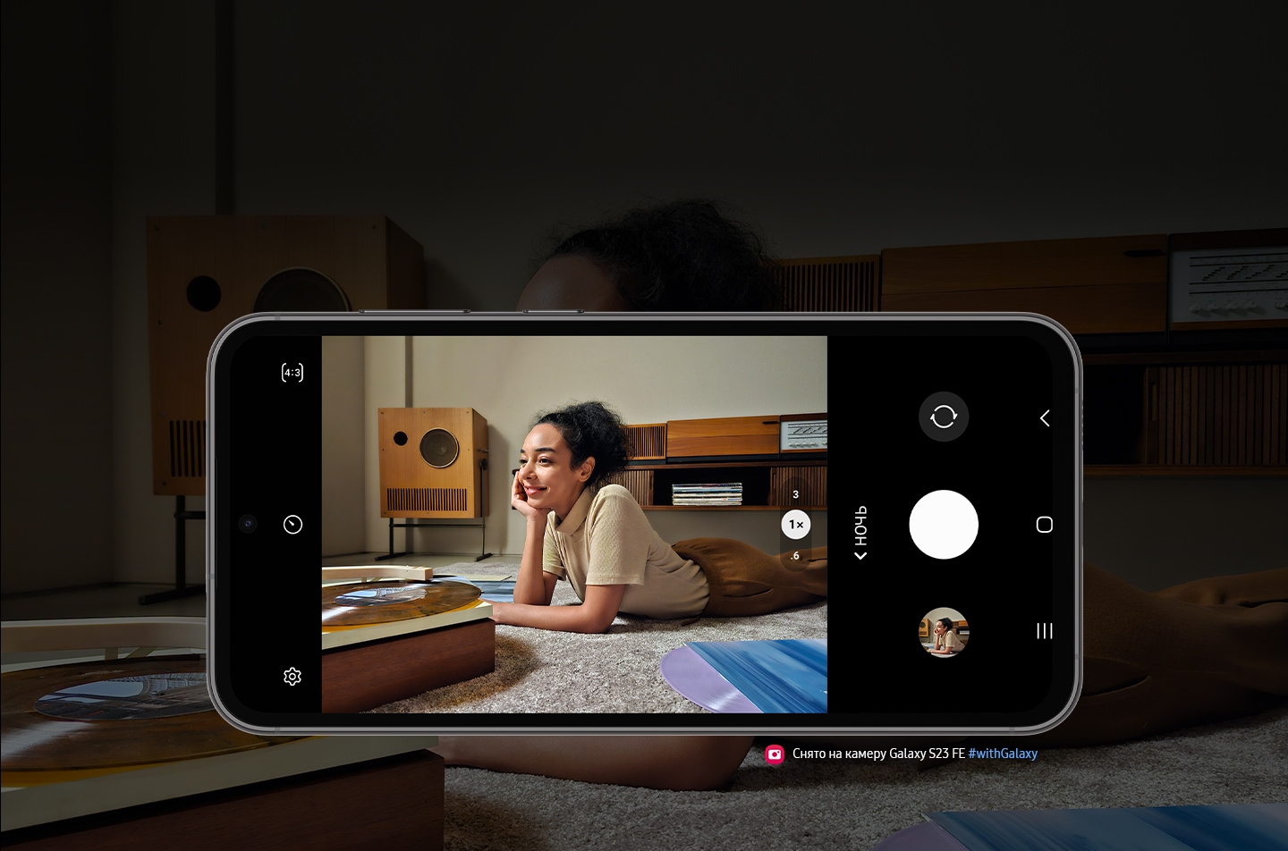 Galaxy S23 FE показан в горизонтальном положении со стороны экрана. На экране четкое изображение женщины, слушающей в полутемной комнате музыку с виниловой пластинки. Текст: Снято на Galaxy S23 FE #withGalaxy.