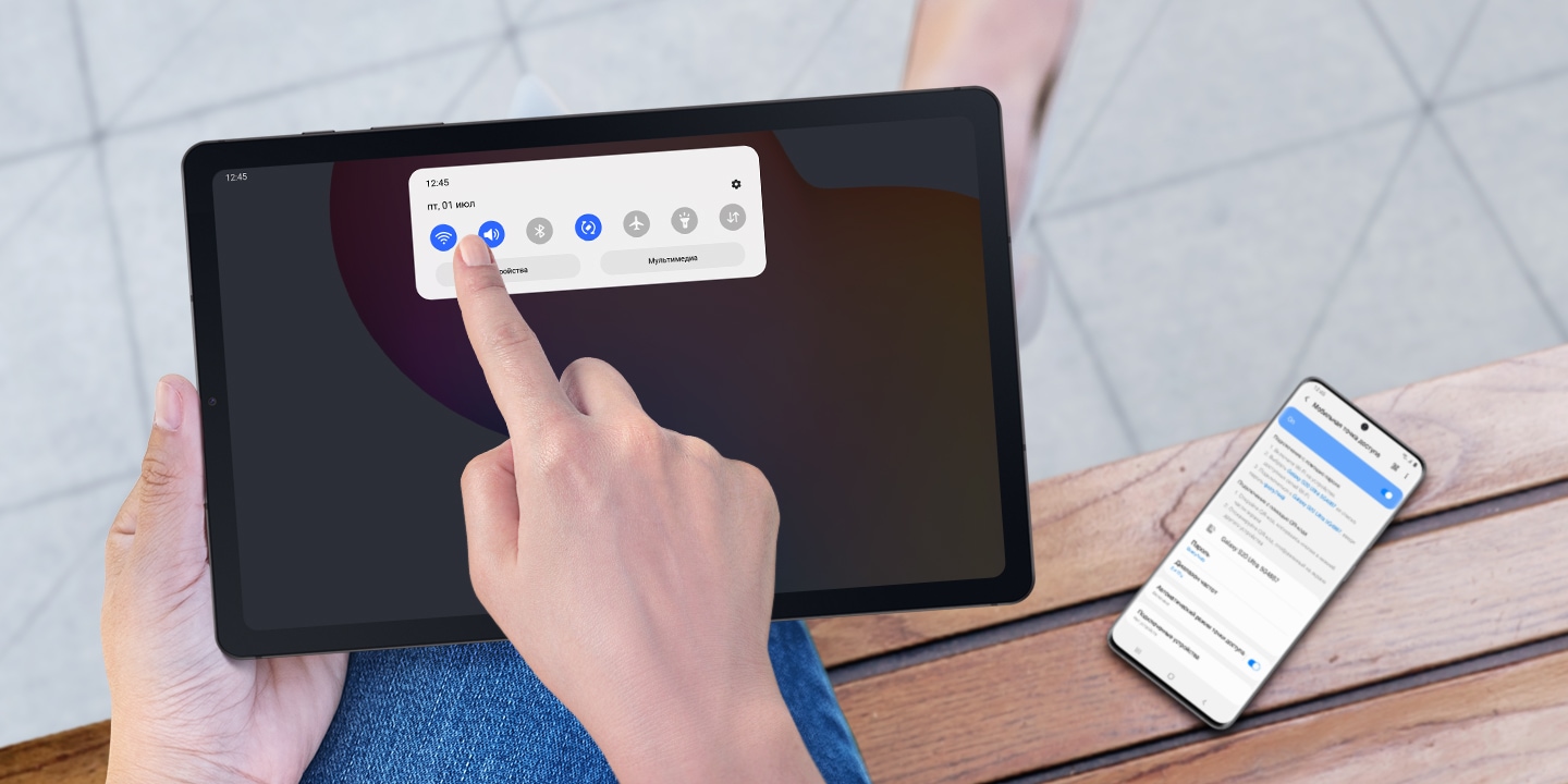 Смартфон Galaxy с включенной функцией мобильной точки доступа лежит на скамейке. Рядом человек держит устройство Galaxy Tab S6 Lite в альбомной ориентации с открытой панелью быстрого доступа и выбранной опцией 