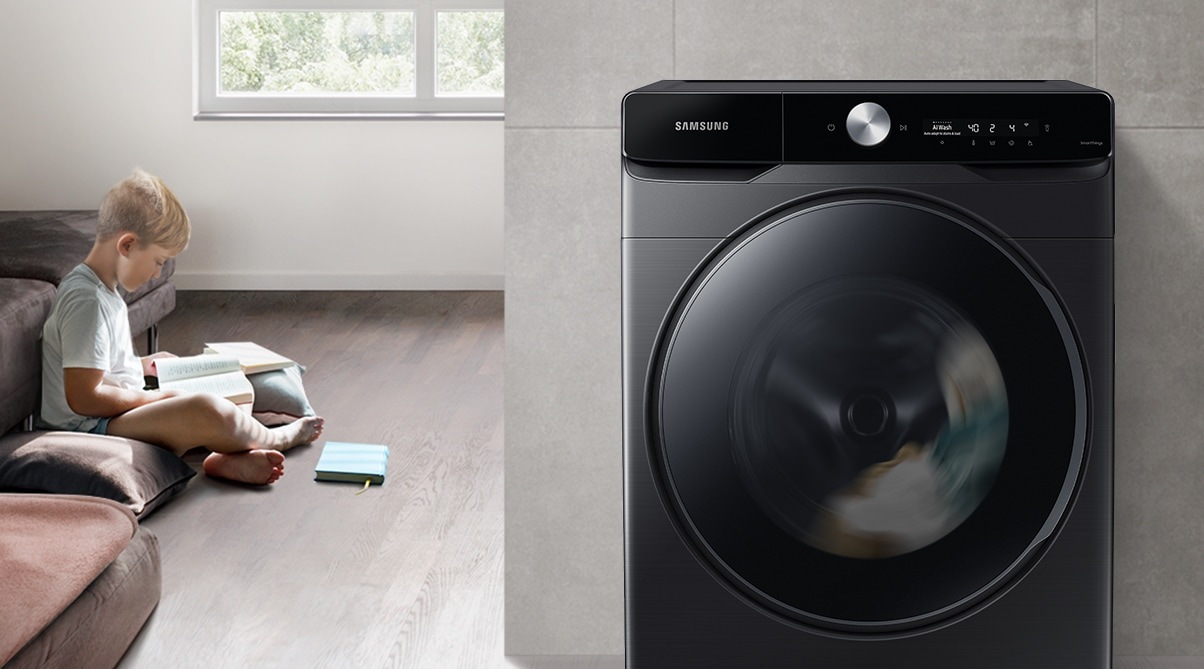 The boy reads the book during the day in the living room, while the washer runs silently next to him.