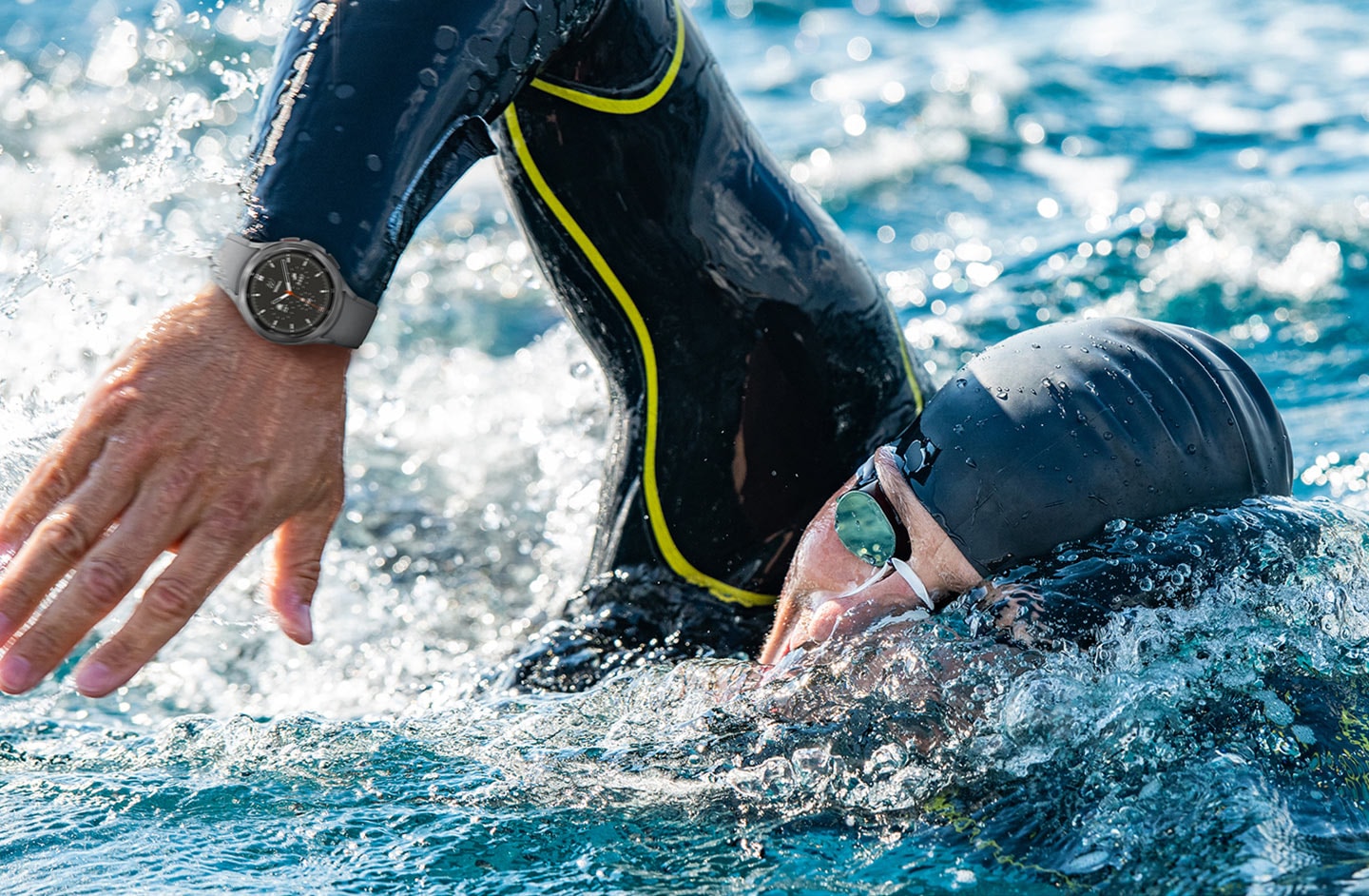 A man is actively swimming and as his right arm is lifted above the water's surface during a swimming stroke, a silver Galaxy Watch4 device attached with a Gray color Ridge band can be seen on his right hand wrist.