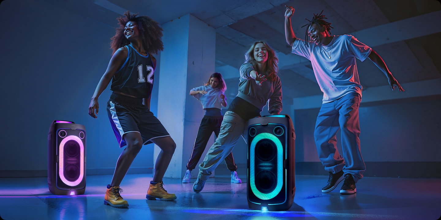 Four people dancing in large room with two Samsung Sound Towers. The dark room is lit up by the speakers' LED party lights.
