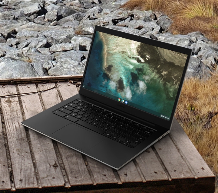 A Galaxy Chromebook Go stands upright, in laptop mode, on a wooden table outdoors.  In the background can be seen an uncountable number of large gray stones and some grass on the ground.