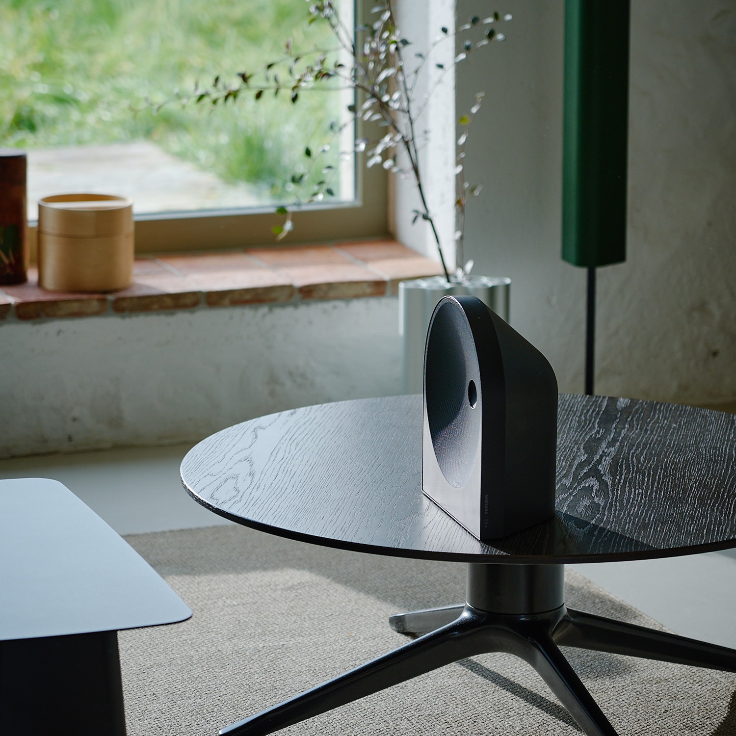 Music Studio 5-högtalare placerad på ett träbord i ett soligt, minimalistiskt vardagsrum. Den svarta, bågformade högtalaren smälter naturligt in i inredningen, tillsammans med en växt.