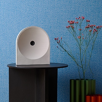 Music Studio 5-högtalare placerad på ett träbord, minimalistiskt vardagsrum med blå väggar. Den svarta, bågformade högtalaren smälter naturligt in i inredningen, tillsammans med en blomma.