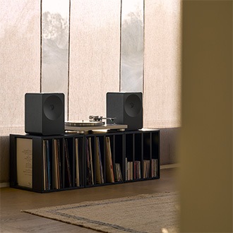 Music Studio 7-högtalare på en trähylla bredvid en grammofon. Högtalaren står bland böcker och inredningsdetaljer och skapar en varm och mysig atmosfär i ett modernt vardagsrum.