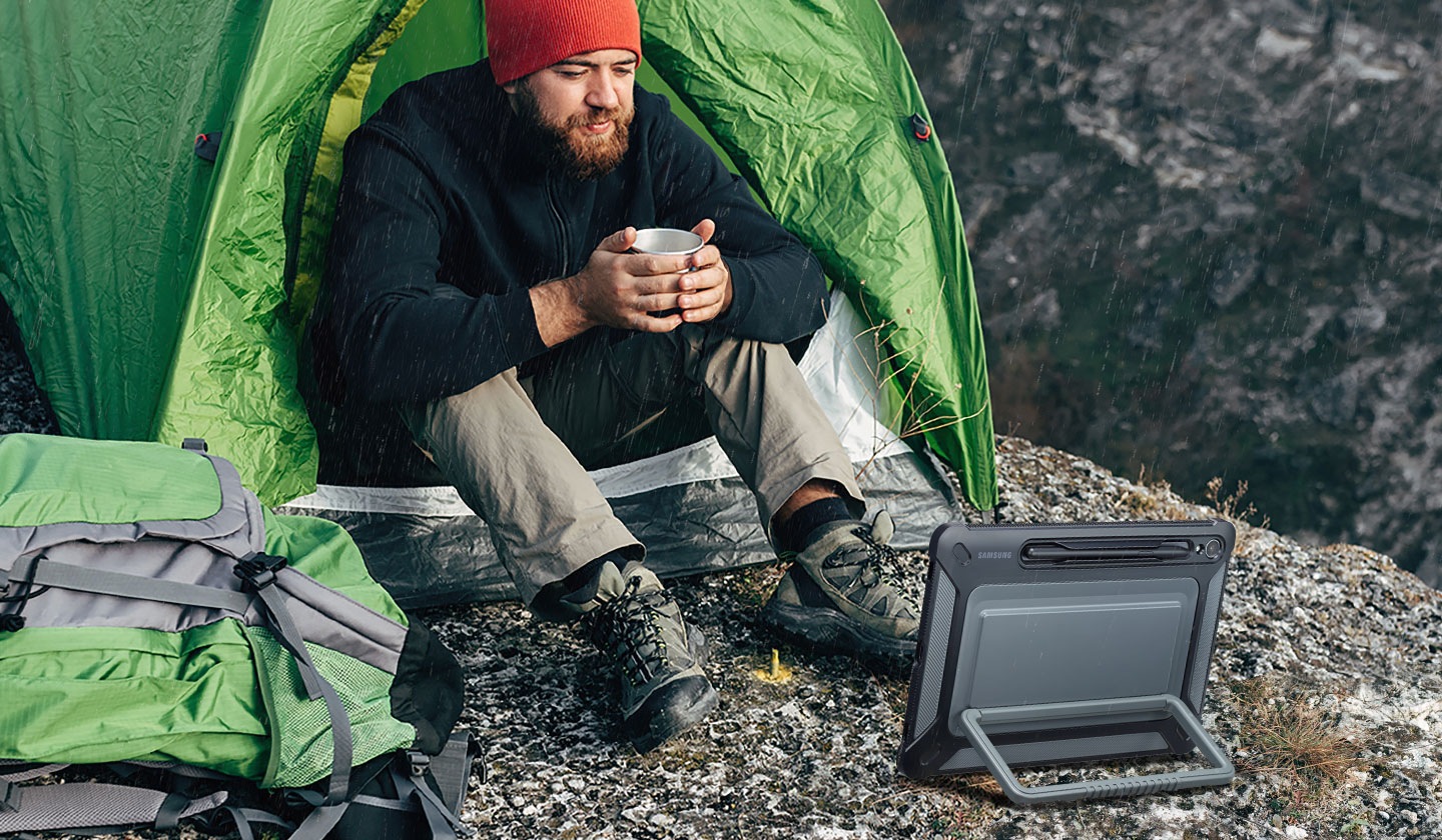 Một người đàn ông cầm cốc đang ngồi trước lều cắm trại trên bề mặt đá, đang nhìn vào thiết bị Galaxy Tab S9 có Vỏ bọc ngoài trời, được đặt nằm ngang bằng chân đế ở mặt sau.