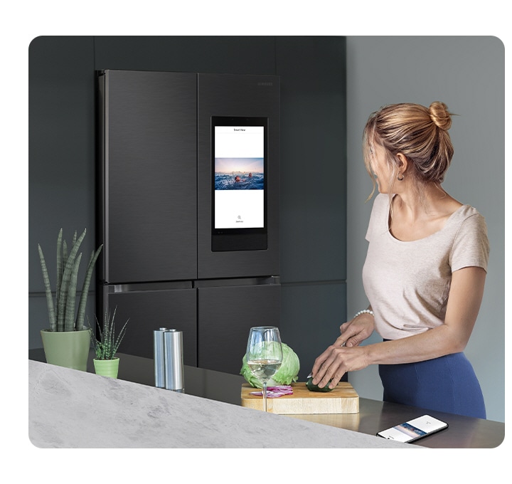 In a stylish kitchen, a woman has a glass of wine while slicing vegetables and watching what’s on the Family Hub’s display.