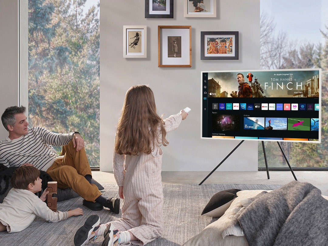 A girl is pointing the remote towards The Frame, which shows the Smart Hub home screen.