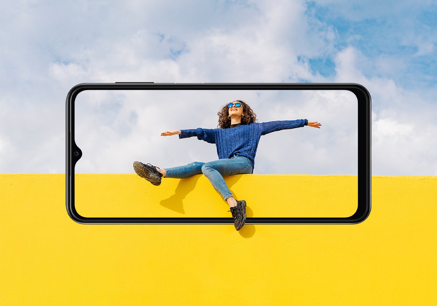 Samsung Galaxy A13 specs and features. Display specs, a scene of a young woman sitting on yellow wall against cloudy sky looking up. Her legs and the wall and sky extend outside of the display.