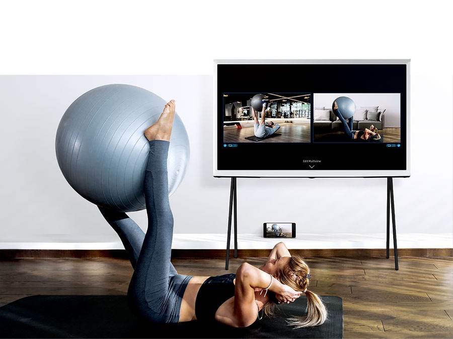 A woman is working out with The Serif's Multi View function to see her reflection and a trainer's demonstration side by side.