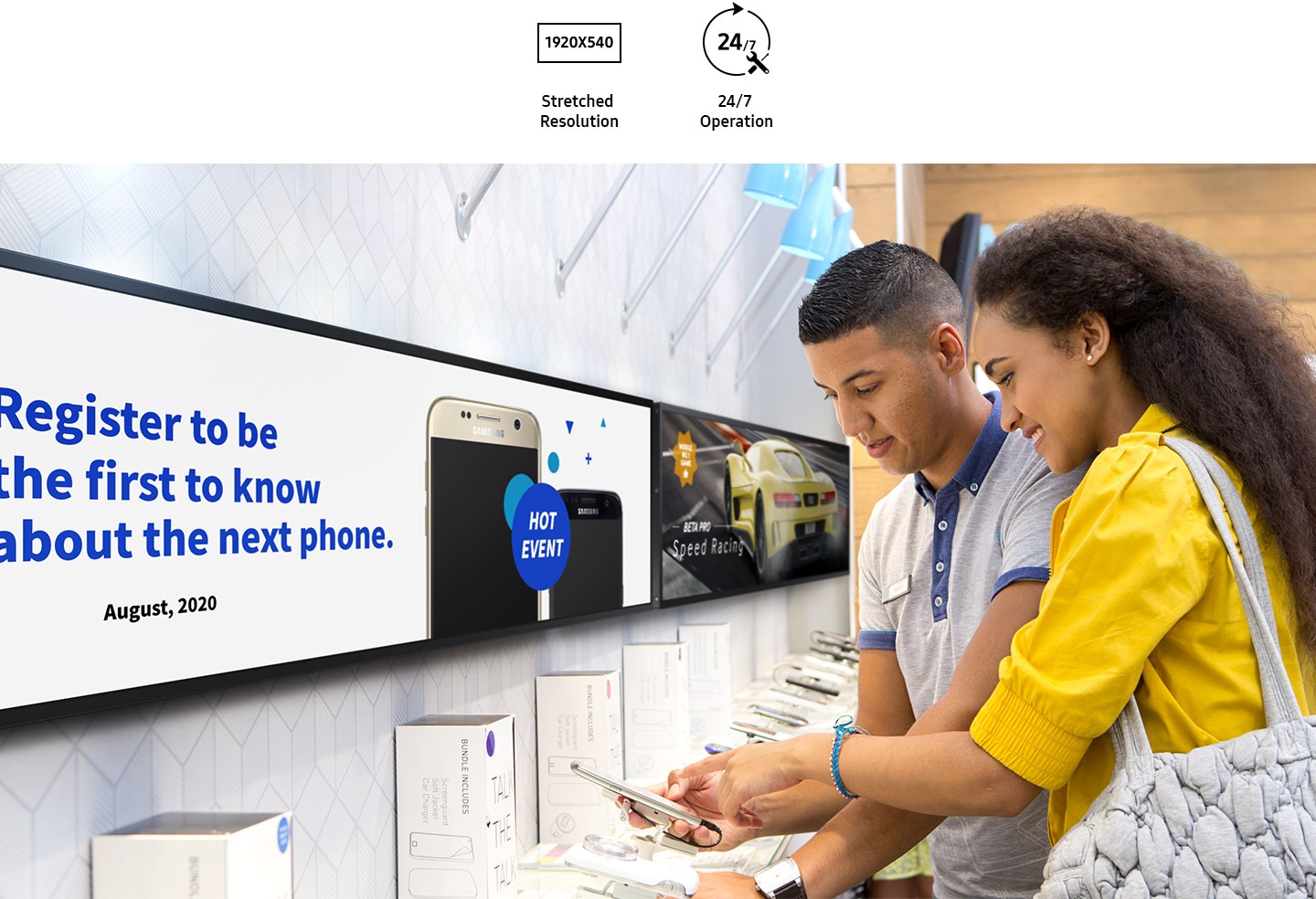 A man and a woman are looking at a cell phone in a cell phone store. Mobile phone advertisements are displayed on the wall-mounted SHR-B. At the top is a 1920X540 stretched resolution and 24/7 operation icon.
