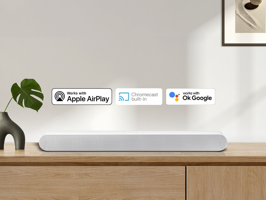 Apple AirPlay logo, Chromecast Built-in logo, and Ok Google logo can be seen along with Samsung S61B soundbar which is sitting on living room cabinet.