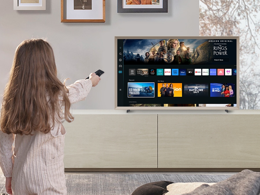 A girl is pointing the remote towards The Frame, which shows the Smart Hub home screen.