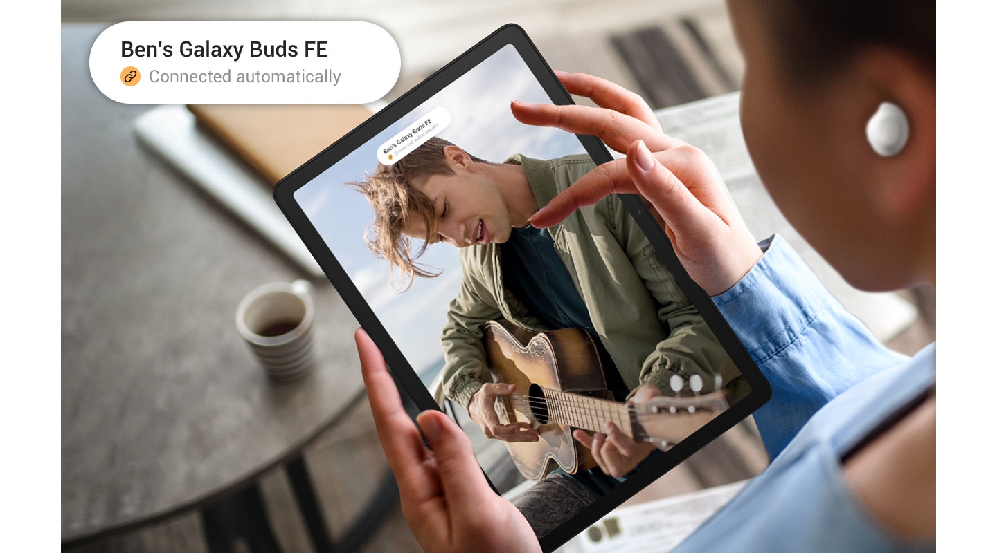 A person is using Galaxy Tab A9+ with Galaxy Buds FE on. On the screen, a man can be seen playing the guitar. A notification is at the top of the screen, and the magnified version of the same notification can be seen in the top left corner. The notification reads Ben's Galaxy Buds FE, and the next line has a link icon with the text Connected automatically.