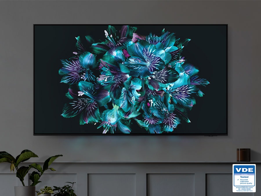 A TV is displaying a colorful flower-like pattern on its screen. The color details of the flower are very vivid. VDE 01/2024 Tested Accurate calibrated picture quality www.VDEiinfo.com ID. 40057838 Tested by VDE Germany.