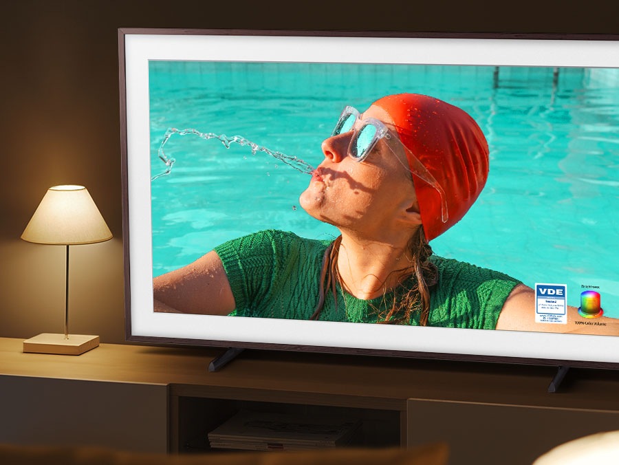 The Frame TV is set in a room with dim lighting, but its screen displays bright, vivid colors powered by Quantum Dot. A woman is spitting out water in the pool. The brightness and 100% color volume powered by Quantum Dot logo is on the lower right side.