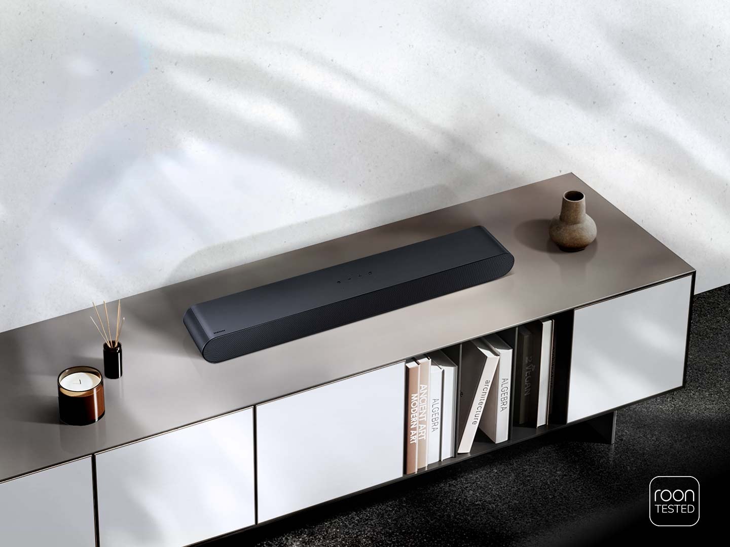 A compact Soundbar is set on top a TV stand, along with other household items. Roon Tested logo.