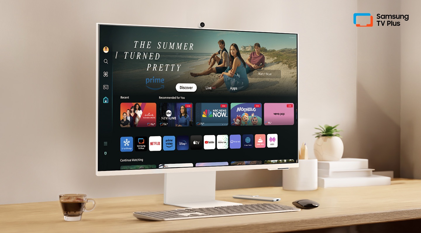 There is a monitor on a desk and its home UI is shown with OTT applications. On the upper right, there's a logo of Samsung TV Plus.