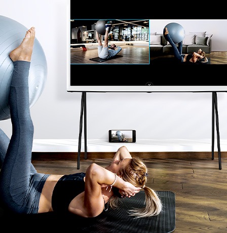 A woman is working out with The Serif's Multi View function to see her image and a trainer's demonstration side-by-side.