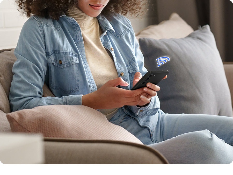 A woman on a couch holds a phone. The laundry recipe tab on the SmartThings app is onscreen. A 56-minute Normal cycle with hot temperature, single rinse, high spin and normal soil levels. End time can be selected on The Laundry Planner tab. A normal cycle with an end time of 11:56 am is selected.