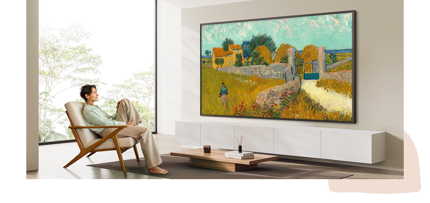 98-inch The Frame TV displays vibrant artwork in a bright living room, as a person relaxes in a lounge chair, enjoying an immersive, gallery-like experience.