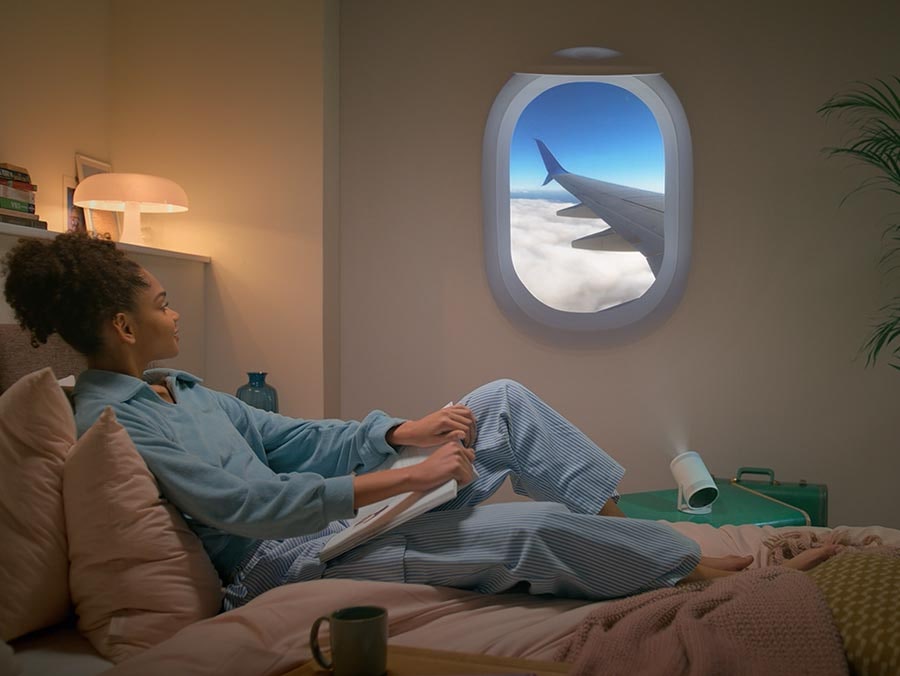 There is a skyline view from an airplane window. The camera zooms out, revealing that it’s actually a wall projection beamed from The Freestyle. A woman is in her bed reading a book, enjoying the simulated flight experience.