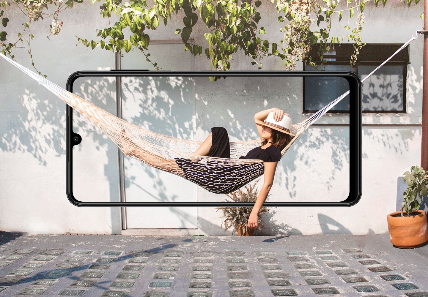 Galaxy A22 je videný spredu, vodorovne. Na obrazovke leží žena v hojdacej sieti, ktorá splýva za obrubou s bokom domu.