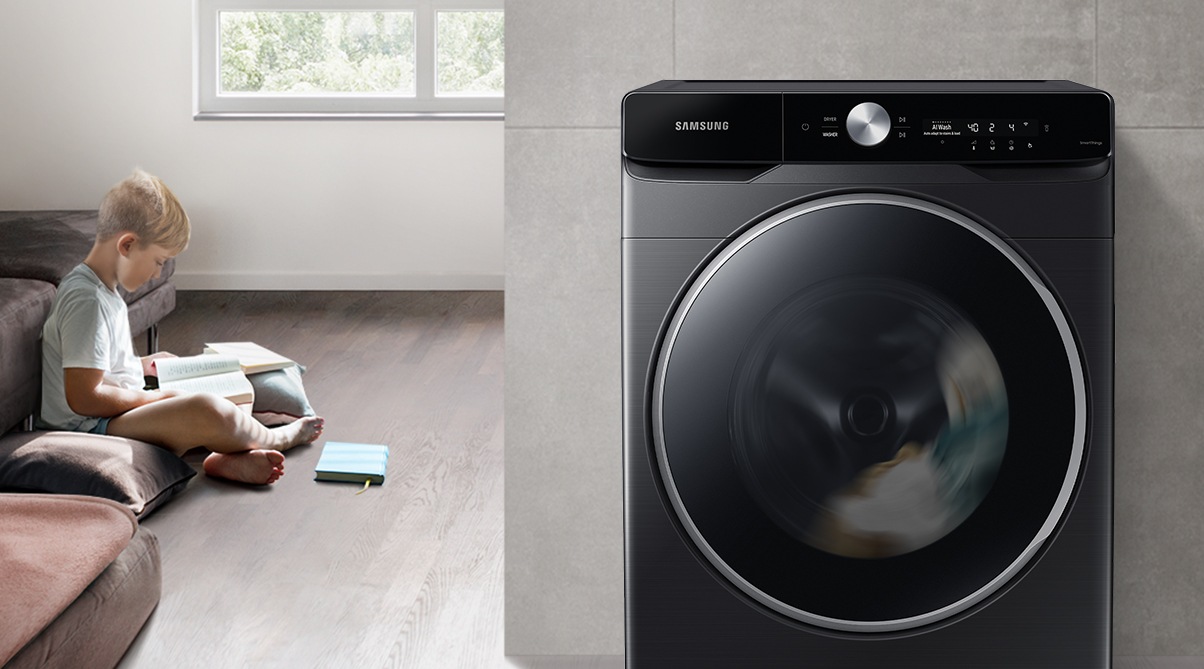 The boy reads the book during the day in the living room, while the washer runs silently next to him.