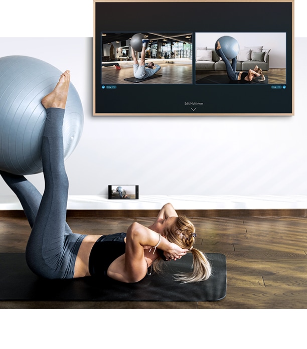 A woman is working out in front of her TV. On the TV screen she can see a screen of herself as well as the screen of the fitness trainer and below the TV her smartphone camera is capturing her workout so she can see it on the TV screen.