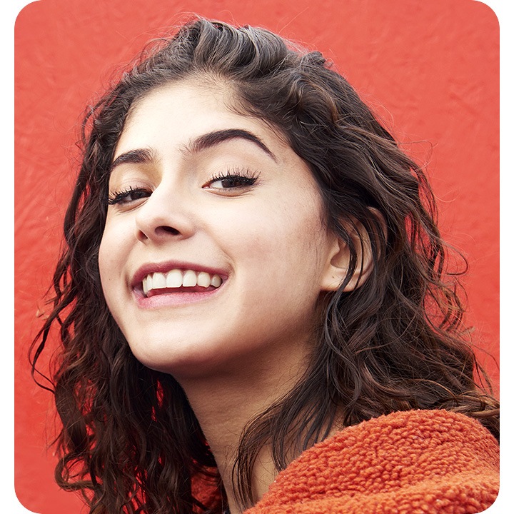 A selfie of a woman standing against a red wall that's gently blurred. She's clear, and seen smiling and looking down into the camera.