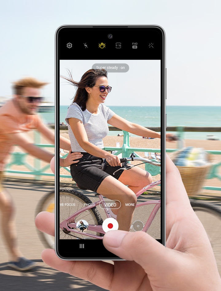 A person on a bike with another person running behind them and pushing. In front of this is a hand holding Galaxy A52s 5G with the camera interface onscreen. The scene in the display is clearer than the scene outside of the display, depicting how Super Steady allows video to be captured smoothly even if the subject is in motion.