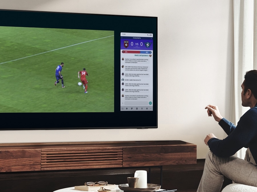 ผู้ชายคนหนึ่งกำลังใช้คุณลักษณะ Multi View ของ QLED เพื่อชมเกมการแข่งขันฟุตบอลและดูข่าวอยู่ในหน้าจอเดียวกันพร้อม ๆ กัน
