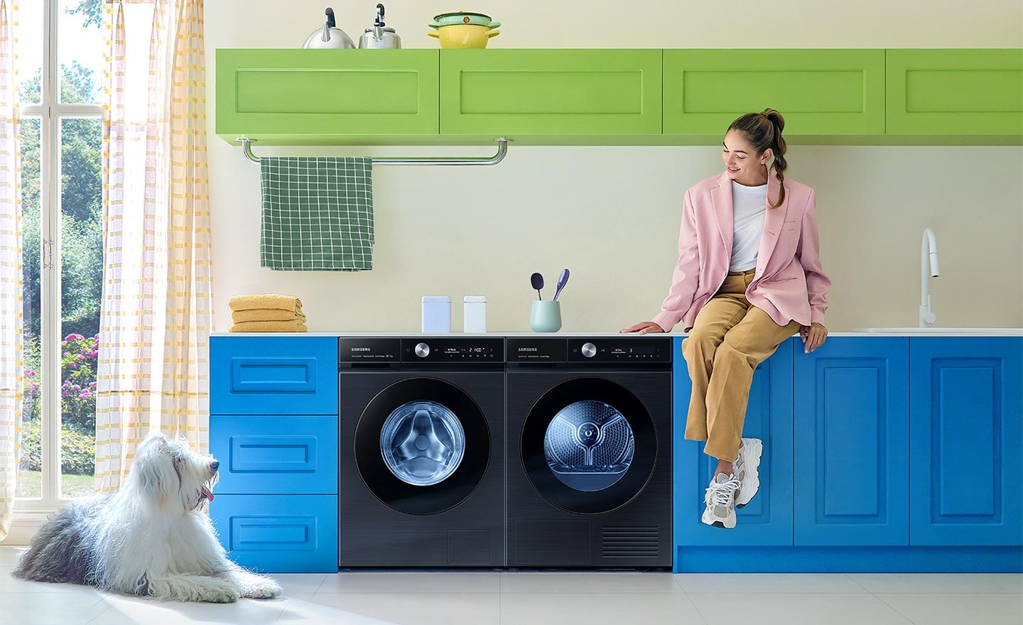 A white Bespoke Grande AI washer and dryer set are built side by side into a green cabinet under a countertop. Towels, detergent, and home accessories are arranged nearby.