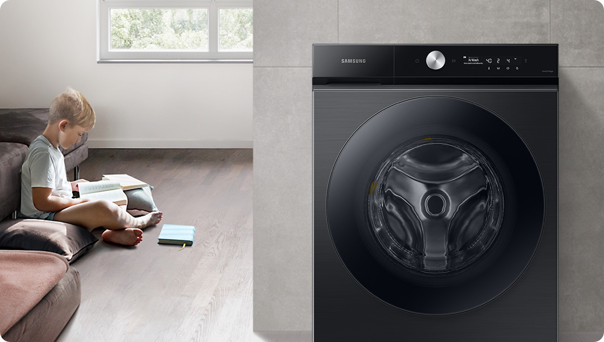 A child sits on the floor in a bright, sunlit room surrounded by books and pillows. To the right is a wall, in front of which is a white Samsung Bespoke Grande AI washer with laundry spinning inside.