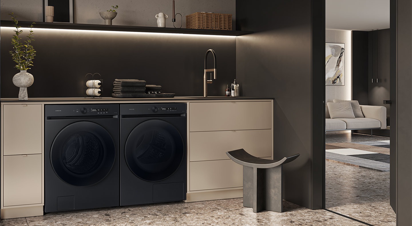 A white Bespoke Grande AI washer and dryer set are built side by side into a green cabinet under a countertop. Towels, detergent, and home accessories are arranged nearby.