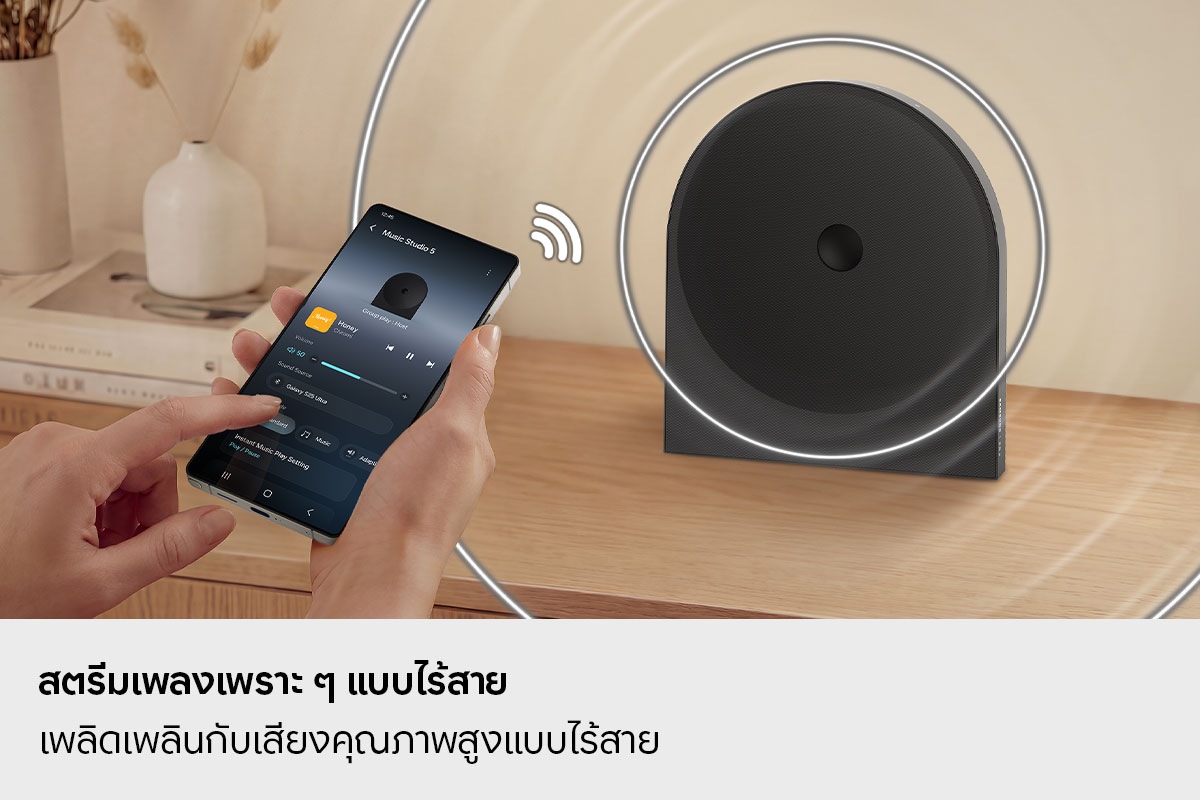บุคคลหนึ่งใช้สมาร์ทโฟนเพื่อสตรีมเพลงแบบไร้สายไปยังลำโพง Music Studio 5 ที่วางอยู่บนโต๊ะ แสดงให้เห็นถึงการเชื่อมต่อ Wi-Fi และ Bluetooth ที่ง่ายดายเพื่อการเล่นเพลงที่ราบรื่น