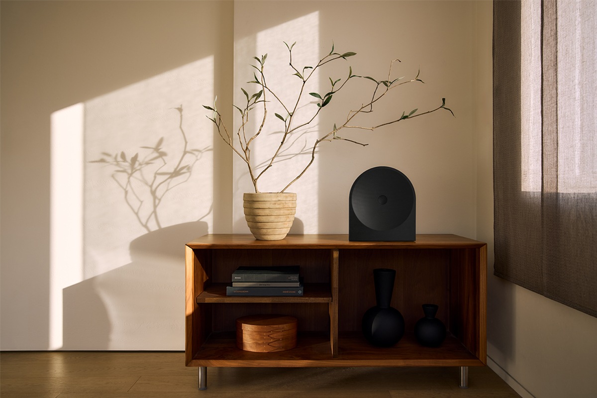 ลำโพง Music Studio 5 วางอยู่บนตู้ไม้ในห้องนั่งเล่นสไตล์มินิมอลที่แสงแดดส่องถึง ลำโพงสีดำทรงโค้งกลมกลืนกับของตกแต่งได้อย่างลงตัว พร้อมกับต้นไม้ที่วางอยู่ข้างๆ