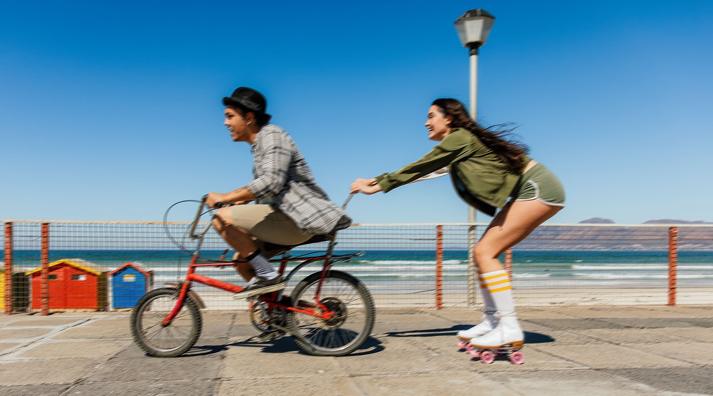 A split-screen with a line in the middle shows a man and a woman near the beach. On the left, which appears slightly blurry, the man is riding a bike. On the right side, which is sharp and clear, the woman is holding onto the man's bike wearing roller skates, highlighting the OIS feature of Galaxy A26 5G.