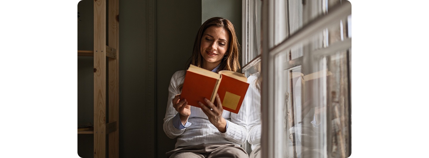 A woman sitting indoors, reading by a window. A line down the screen splits it into two sections, comparing a low-light shot on the left with a bright-light shot on the right.